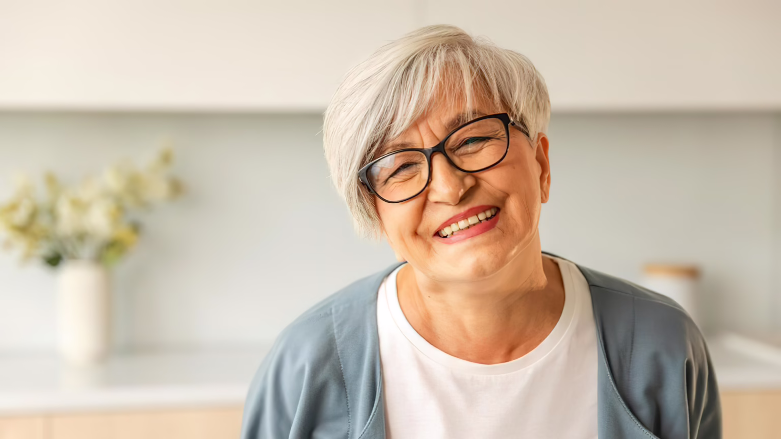 Smiling older woman with short gray hair and black glasses, looking at the camera in a bright room.