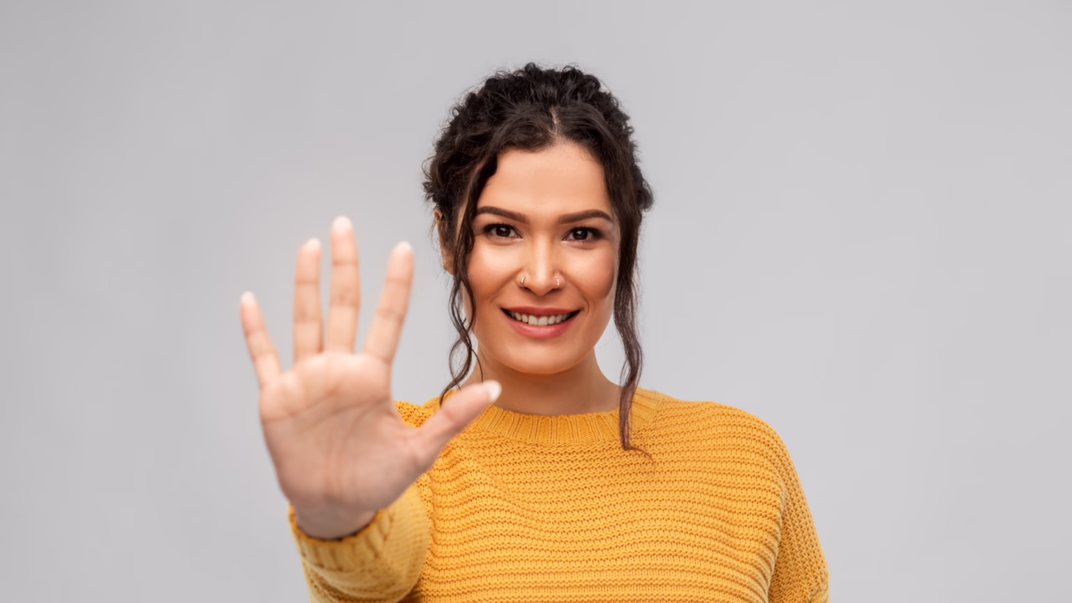 Smiling woman with brown hair in an orange knit sweater raises her hand toward the camera as a friendly hello gesture.