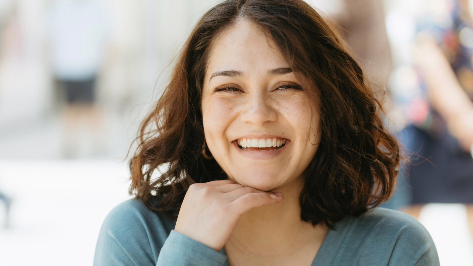 Close-up of a smiling woman with brown hair wearing a teal top, hands lightly resting under her chin.