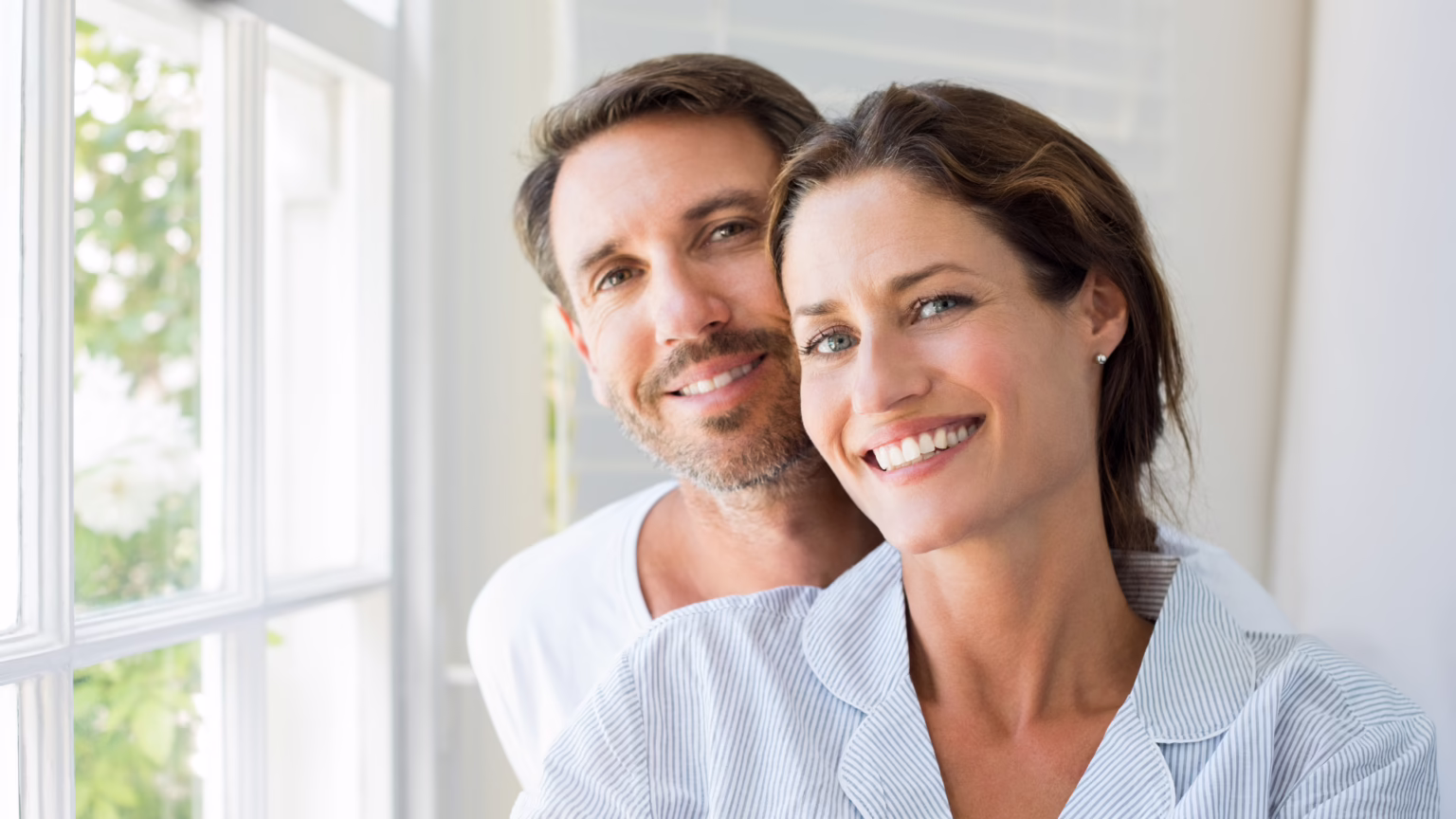 Close-up of a smiling woman in a striped shirt with a man behind her, posing by a sunlit window in a bright room.